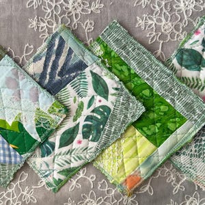 May include: A set of four square quilted coasters with a variety of green and white botanical fabric patterns. Each coaster has a different design, including leaf prints, checkered patterns, and abstract designs. The edges are finished with a decorative trim.