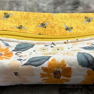 May include: A rectangular, zippered pouch with a yellow bee-themed top and a floral print bottom. The floral pattern features orange and yellow flowers with green leaves on a white background. A yellow handle is attached to the side.