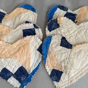 May include: A set of six heart-shaped quilted fabric coasters featuring a patchwork design in cream, peach, and dark blue. Each coaster has a scalloped edge and visible stitching patterns that follow the heart shape across the fabric surface.