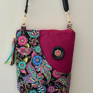 May include: A crossbody bag with a black strap and gold-colored hardware. The bag features a vibrant floral pattern on a black background, with pink, blue, and teal flowers. A solid magenta panel is on the front, with a large black button with a flower design.