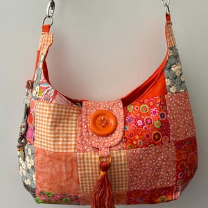 May include: A patchwork hobo bag featuring various orange, pink, and grey floral and gingham fabric squares. The bag has a central flap secured by a large orange button, an orange tassel, and a brown strap with silver hardware and a charm.