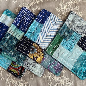 May include: A collection of handmade, square coasters with a patchwork design. The coasters feature various shades of blue, teal, and green fabrics, stitched together with white thread. Some patches have floral patterns, while others have geometric designs.