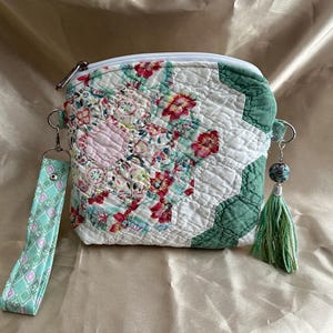 May include: A quilted zipper pouch featuring a hexagonal patchwork pattern with floral prints in shades of pink, red, and white, accented by solid green fabric. It includes a detachable mint green wrist strap with a diamond pattern and a green tassel.