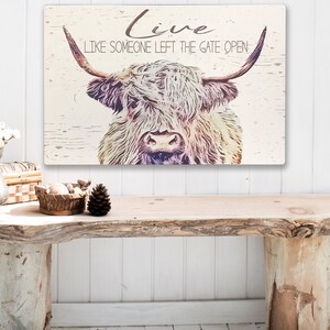 Op de afbeelding: Rustieke wandkunst met een Schotse Hooglander met lange hoorns en een ruige vacht. De tekst "Live Like Someone Left The Gate Open" staat in een scriptletter. Het kunstwerk heeft een verweerde, vintage uitstraling op een lichte houten achtergrond.