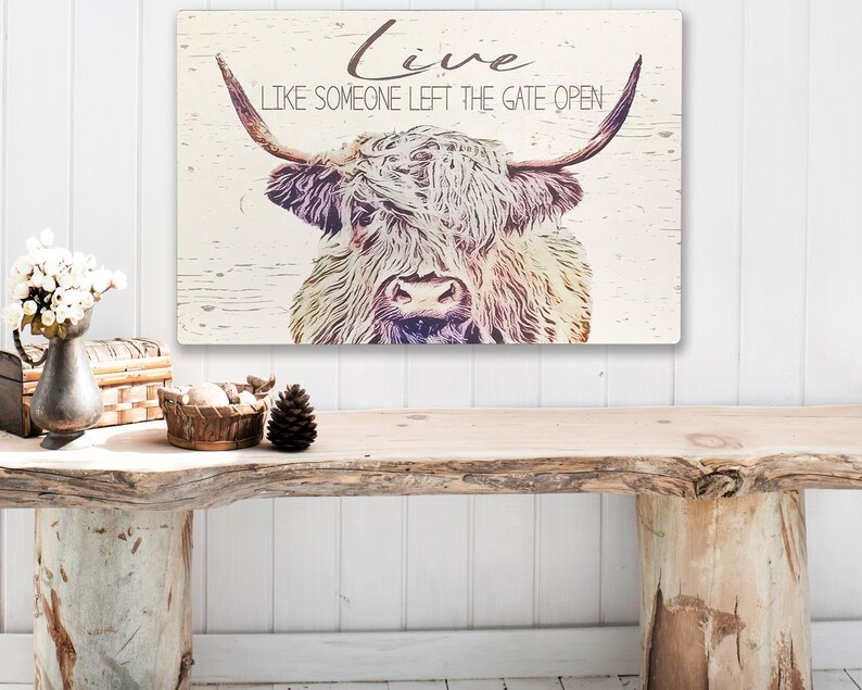 Op de afbeelding: Rustieke wandkunst met een Schotse Hooglander met lange hoorns en een ruige vacht. De tekst "Live Like Someone Left The Gate Open" staat in een scriptletter. Het kunstwerk heeft een verweerde, vintage uitstraling op een lichte houten achtergrond.