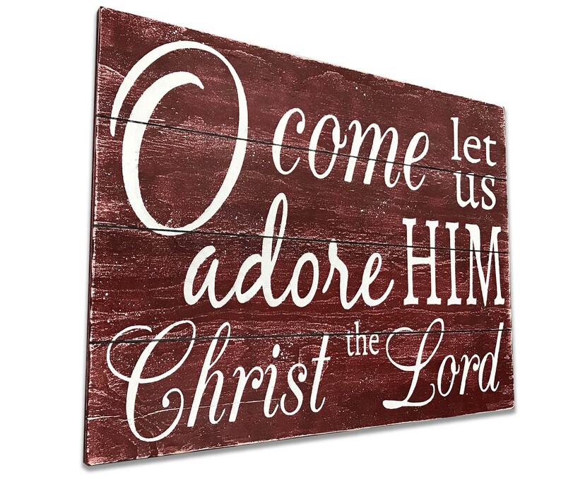 May include: A rustic wooden sign with a red background and white lettering. The sign reads "Come let us adore HIM Christ the Lord".