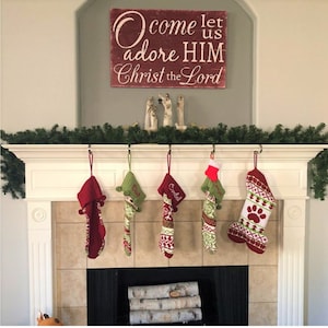 May include: A red wooden sign with white lettering that reads "Come let us adore HIM Christ the Lord".  The sign is hanging above a white fireplace mantel with five stockings hanging from hooks. The stockings are red and green with white accents and are personalized with names.