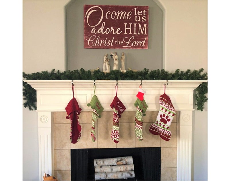 May include: A red wooden sign with white lettering that reads "Come let us adore HIM Christ the Lord".  The sign is hanging above a white fireplace mantel with five stockings hanging from hooks. The stockings are red and green with white accents and are personalized with names.