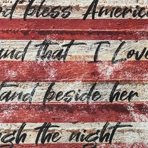 May include: A distressed wood sign with the words "God bless America, land that I love, stand beside her through the night" painted in black on a red, white, and blue American flag background.