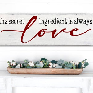 May include: White wooden sign with black text that reads "the secret ingredient is always" and a red cursive word "love".