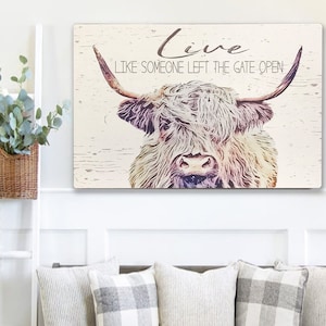 Op de afbeelding: Een rechthoekig houten bord met een illustratie van een Schotse Hooglander en de tekst "Live Like Someone Left The Gate Open". De koe heeft lang, ruig haar en grote hoorns. De achtergrond is licht, verweerd hout.