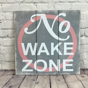 May include: A gray wooden sign with a pink circle around the words "No Wake Zone".