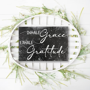 May include: Black chalkboard sign with white text that reads "Inhale Grace Exhale Gratitude" surrounded by white flowers and greenery.
