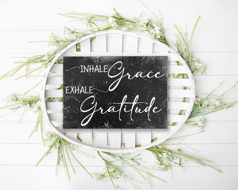 May include: Black chalkboard sign with white text that reads "Inhale Grace Exhale Gratitude" surrounded by white flowers and greenery.