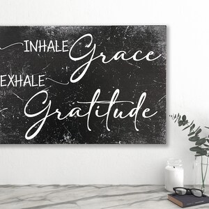 May include: Black and white sign with a distressed texture. The sign reads "INHALE grace EXHALE gratitude" in a white cursive font.