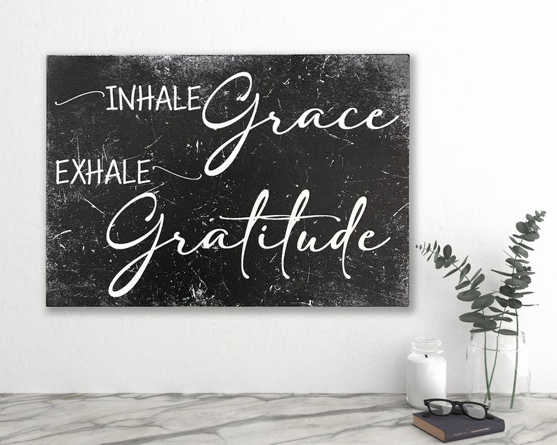 May include: Black and white sign with a distressed texture. The sign reads "INHALE grace EXHALE gratitude" in a white cursive font.