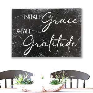 May include: Black and white sign with the text "INHALE grace EXHALE gratitude" on a distressed background. The sign is hanging on a white wall above a table with two chairs.