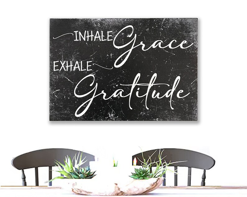 May include: Black and white sign with the text "INHALE grace EXHALE gratitude" on a distressed background. The sign is hanging on a white wall above a table with two chairs.