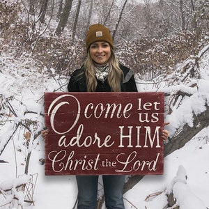 May include: A woman in a winter setting holds a red wooden sign with white lettering that reads "O come let us adore HIM Christ the Lord".