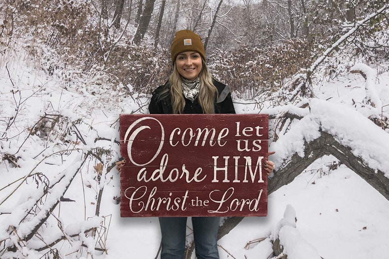 May include: A woman in a winter setting holds a red wooden sign with white lettering that reads "O come let us adore HIM Christ the Lord".