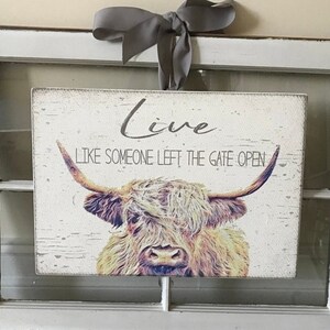 Op de afbeelding: Rechthoekig houten bord met een verweerde afwerking, met een Highland koe illustratie en de tekst "Live Like Someone Left The Gate Open". Een grijs lint is bovenaan vastgebonden om op te hangen.