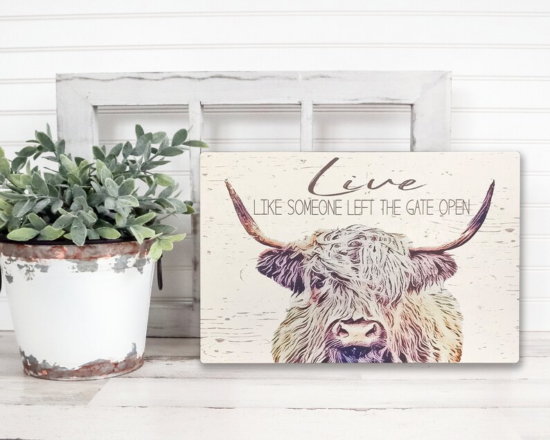 Op de afbeelding: Rechthoekig houten bord met een verweerde afwerking met een illustratie van een Schotse Hooglander. Het bord heeft de tekst "Live Like Someone Left The Gate Open" in een handgeschreven lettertype. Een witte emmer met groen staat op de achtergrond.