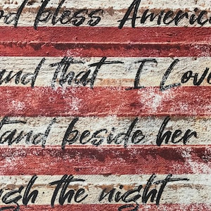 May include: A distressed wood sign with the words "God bless America, land that I love, stand beside her through the night" painted in black on a red, white, and blue American flag background.