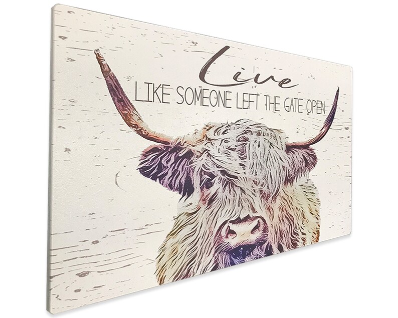 Op de afbeelding: Een rechthoekige wandkunstprint met een Schotse Hooglander met lange hoorns en ruige vacht. Het kunstwerk heeft een verweerde, vintage uitstraling met de woorden "Live Like Someone Left The Gate Open" in een scriptlettertype.