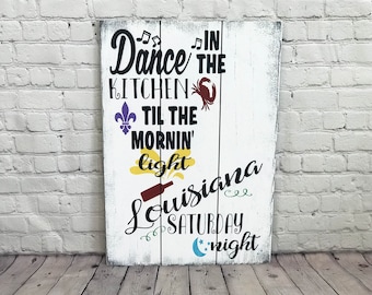 Puede incluir: Señal de madera blanca con texto negro que dice "Dance in the Kitchen til the mornin' light Louisiana Saturday night". La señal tiene un cangrejo rojo, una flor de lis morada, un sol amarillo y una luna creciente azul con estrellas.