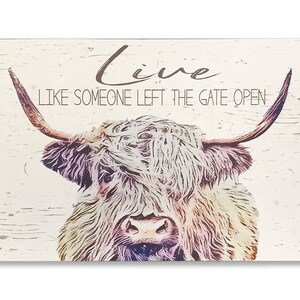 Op de afbeelding: Rechthoekig houten bord met een illustratie van een Schotse Hooglander. De tekst "Live Like Someone Left The Gate Open" staat op het bord. De koe heeft lange hoorns en een ruige vacht. Lichte houtnerf achtergrond.