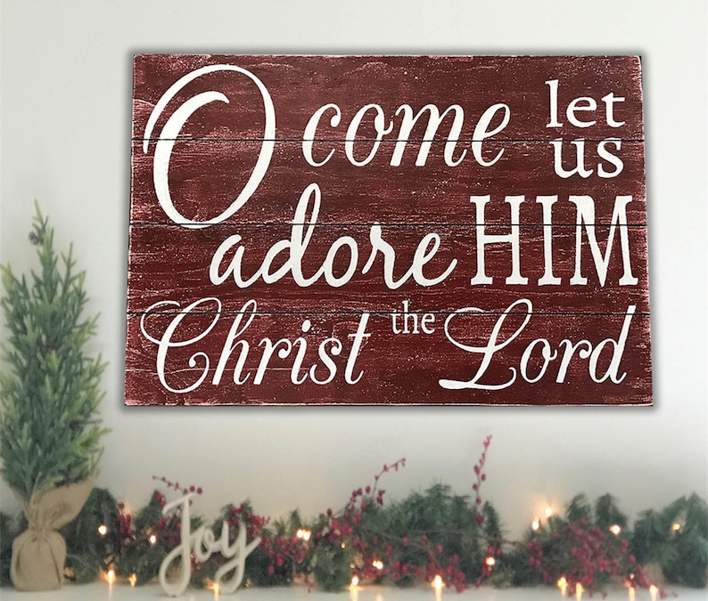 May include: Red wooden sign with white lettering that reads "O come let us adore HIM Christ the Lord".