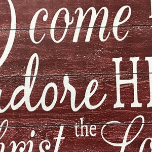 May include: Red wooden sign with white lettering that reads "O come let us adore Him, Christ the Lord".