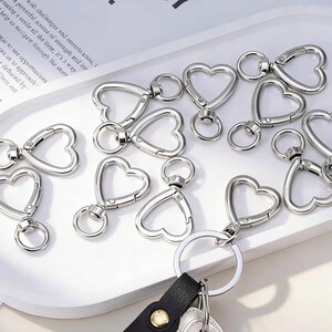 May include: A collection of silver-toned metal heart-shaped swivel lobster clasps. Each clasp features a heart-shaped frame with a spring-loaded gate and a small circular ring attached to the base for connecting to keychains, jewellery, or accessories.