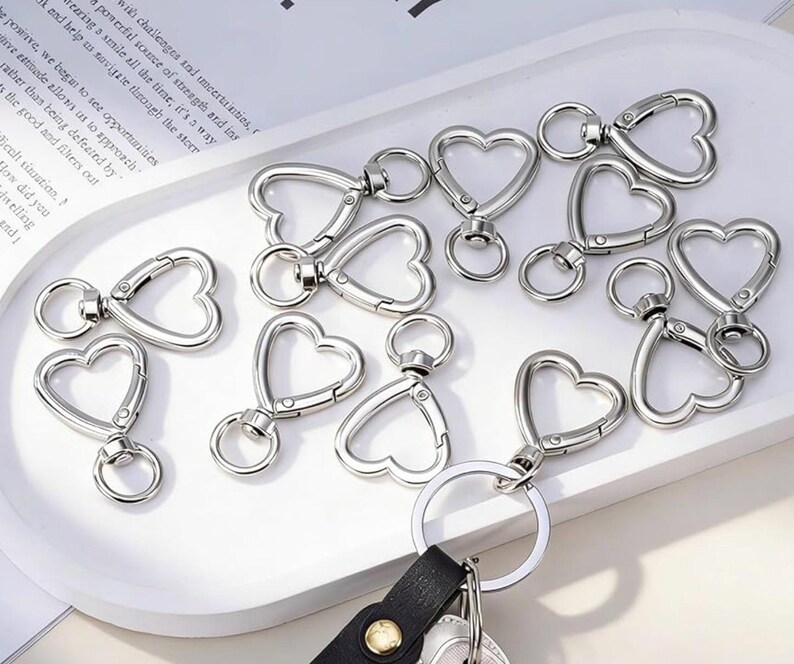 May include: A collection of silver-toned metal heart-shaped swivel lobster clasps. Each clasp features a heart-shaped frame with a spring-loaded gate and a small circular ring attached to the base for connecting to keychains, jewellery, or accessories.