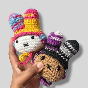 May include: Two small, round, crocheted plush toys with long ears. One toy has a white face with black eyes and a cross-stitched mouth, wearing a yellow and pink patterned hat. The other has a tan face with black eyes and a black and purple hat.