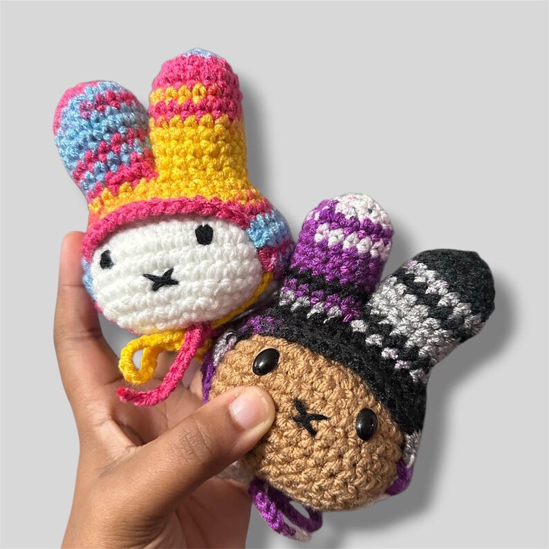 May include: Two small, round, crocheted plush toys with long ears. One toy has a white face with black eyes and a cross-stitched mouth, wearing a yellow and pink patterned hat. The other has a tan face with black eyes and a black and purple hat.