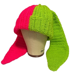 May include: A crocheted hat with a pink and green color scheme. The hat has two long tails that hang down the back.