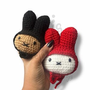 May include: Two small crochet plush toys featuring rabbit-like ears. One toy has a tan face with a black hood, while the other has a white face with a red hood. Both have black button eyes and a stitched black cross for a mouth, crafted in yarn.