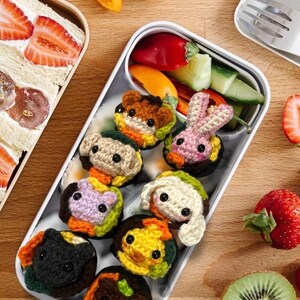 K&ouml;nnte beinhalten: Eine wei&szlig;e Lunchbox mit bunten, geh&auml;kelten Sandwiches in Tierform, zusammen mit frischem Gem&uuml;se und Obst. Die Sandwiches zeigen verschiedene Tiergesichter, darunter ein B&auml;r, ein Hase und ein Hund. Erdbeeren, Kiwi und Paprika sind ebenfalls zu sehen.