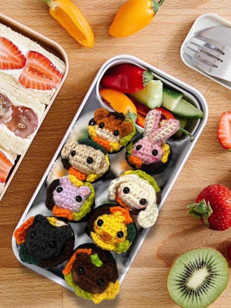K&ouml;nnte beinhalten: Eine wei&szlig;e Lunchbox mit bunten, geh&auml;kelten Sandwiches in Tierform, zusammen mit frischem Gem&uuml;se und Obst. Die Sandwiches zeigen verschiedene Tiergesichter, darunter ein B&auml;r, ein Hase und ein Hund. Erdbeeren, Kiwi und Paprika sind ebenfalls zu sehen.
