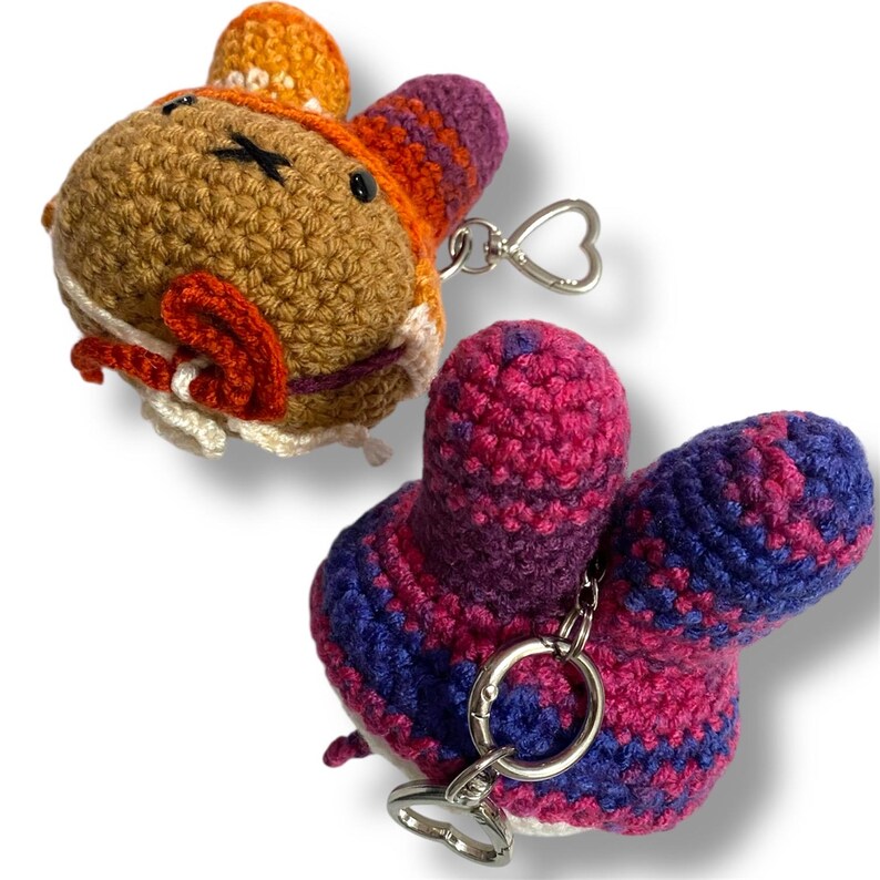 May include: Two crochet keychains featuring rounded shapes with long ears. One is tan with an orange and purple hat and white accents. The other is a mix of pink and blue yarn. Both include a silver metal keyring with a heart-shaped clasp.