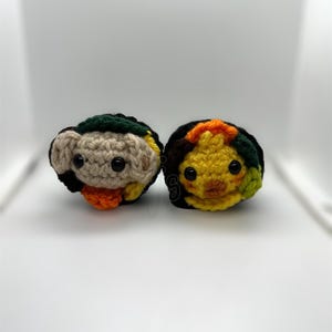 K&ouml;nnte beinhalten: Zwei handgefertigte Amigurumi-Figuren aus H&auml;kelarbeit. Eine ist hellbraun mit schwarzen, gr&uuml;nen und orangefarbenen Akzenten. Die andere ist gelb mit schwarzen, orangefarbenen und gr&uuml;nen Akzenten. Beide haben schwarze Knopfaugen und sind auf wei&szlig;em Hintergrund.
