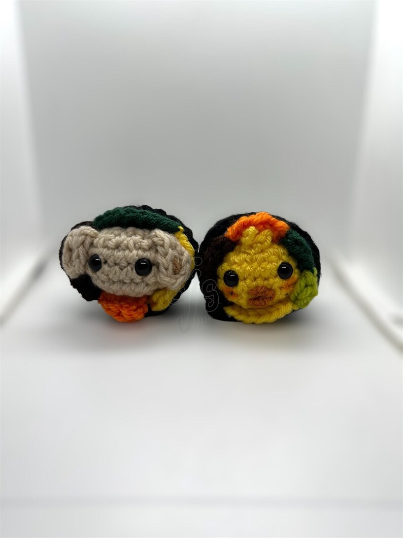 K&ouml;nnte beinhalten: Zwei handgefertigte Amigurumi-Figuren aus H&auml;kelarbeit. Eine ist hellbraun mit schwarzen, gr&uuml;nen und orangefarbenen Akzenten. Die andere ist gelb mit schwarzen, orangefarbenen und gr&uuml;nen Akzenten. Beide haben schwarze Knopfaugen und sind auf wei&szlig;em Hintergrund.