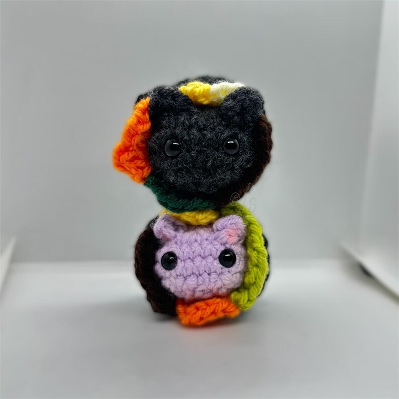 K&ouml;nnte beinhalten: Ein handgefertigtes H&auml;keltierspielzeug in Schwarz, Orange, Gr&uuml;n, Gelb und Lila. Die Katze hat gro&szlig;e schwarze Augen und ist aus weichem Garn gefertigt. Das Spielzeug ist eine einzigartige, farbenfrohe und skurrile Kreation.