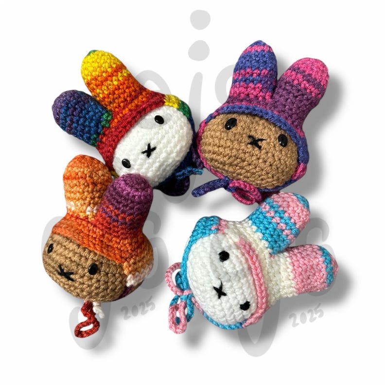 May include: Four small, handmade crochet rabbit plushies with long ears. One has a white face and rainbow-coloured ears, one is brown with purple and pink ears, one is brown with orange and purple ears, and one is white with blue and pink ears.