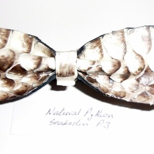Può includere: Un papillon realizzato in pelle di serpente pitone naturale. Il papillon presenta un motivo a scaglie color crema e marrone. Il testo "Natural Python Snakeskin P3" è scritto a mano su una carta bianca sotto il papillon.