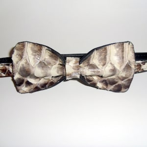 May include: A bow tie made of reptile skin, featuring a pattern of beige, brown, and black scales. The bow tie has a black leather backing and a matching adjustable strap. The accessory is designed to be worn around the neck.