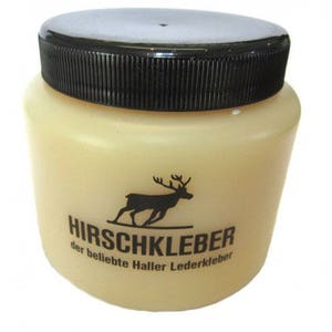 Puede incluir: Un tarro de pegamento para cuero Hirschkleber con tapa negra. El tarro es de color amarillo claro con una silueta negra de un ciervo y el nombre de la marca impreso en negro. El texto "der beliebte Haller Lederkleber" también está impreso.