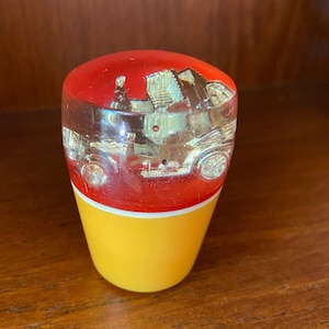 Può includere: Un pomello del cambio in vetro trasparente con una base rossa e gialla. Il pomello presenta un'auto d'epoca all'interno.