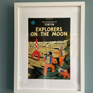 May include: A framed print of a comic book cover titled "Explorers on the Moon" featuring astronauts in orange suits, a red and white rocket, and a dog on a lunar landscape. The print is in a white frame.